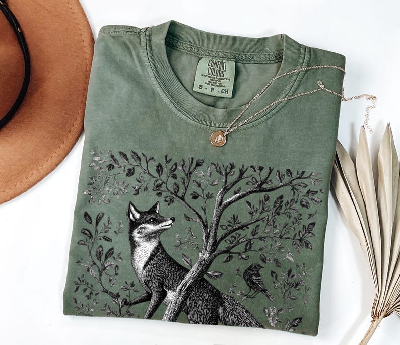 Peut inclure: Un t-shirt vert avec une illustration en noir et blanc d'un renard dans un arbre. Le t-shirt porte le texte "COMFORT COLORS" et "S - P - CH" sur l'&eacute;tiquette.
