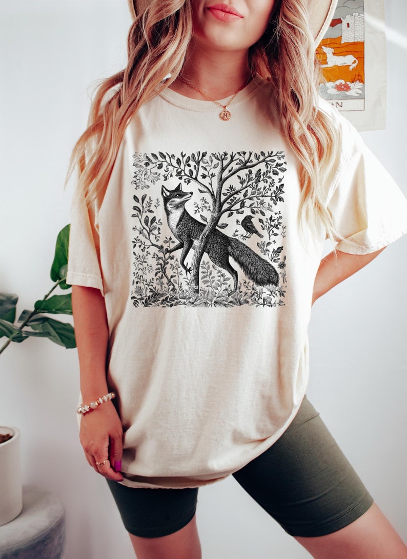 Peut inclure: Un t-shirt cr&egrave;me avec une illustration en noir et blanc d'un renard dans un d&eacute;cor de for&ecirc;t. Le renard est debout sur ses pattes arri&egrave;re et regarde vers la gauche. Un oiseau est perch&eacute; sur une branche au-dessus du renard.
