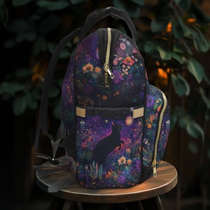 May include: A black backpack with a floral and starry night design. The backpack has a large zippered pocket on the front and a smaller pocket on the side. The backpack is sitting on a wooden stool.