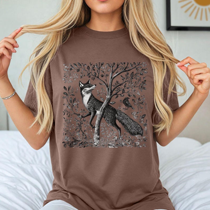 Peut inclure: Un t-shirt marron avec une illustration vintage d'un renard debout dans une for&ecirc;t avec un oiseau perch&eacute; sur une branche.