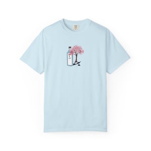 Op de afbeelding: Lichtblauw T-shirt met een afbeelding van een gin-fles en een kersenbloesemboom. De gin-fles is blauw met een wit etiket. De kersenbloesemboom heeft roze bloemen en een zwarte stam. Het shirt is lichtblauw.