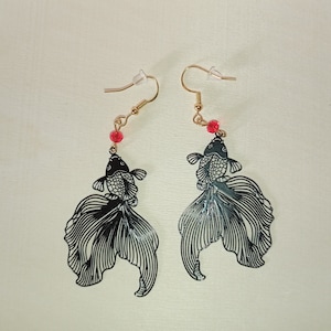 May include: A pair of black dangle earrings featuring a detailed silhouette of a fish with a red bead accent. The earrings are made of metal and have a delicate, feminine design.