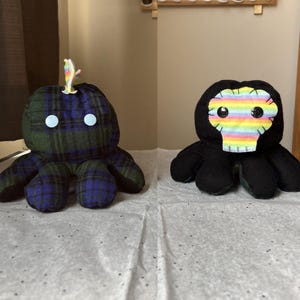 Könnte beinhalten: Zwei Plüsch-Kraken. Einer ist schwarz mit einem Regenbogen-Schädelgesicht und der andere ist grün-blau kariert mit blauen Augen.