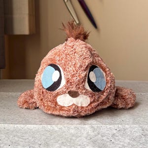 May include: A cartoon animal plush toy with a brown body, large blue eyes, and a tuft of brown hair. The toy has a white muzzle and small arms. The toy is on a gray surface.