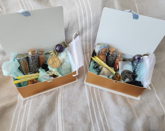 Puede incluir: Dos cajas abiertas con tapas doradas, llenas de varios artículos, incluyendo pequeñas botellas, cristales, velas y un collar. Las cajas están forradas con papel de seda azul y tul blanco.
