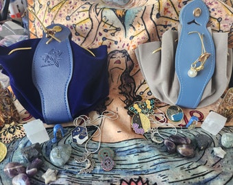 Puede incluir: Dos bolsas de terciopelo azul con detalles dorados y una correa de cuero. Las bolsas están llenas de cristales, amuletos y otros objetos mágicos. Las bolsas están sobre un tapiz con la imagen de una mujer.