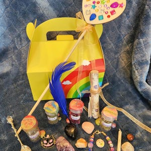 May include: A yellow gift box with a rainbow design, a heart-shaped wand adorned with gems, and various mystical items. Includes jars of herbs, a blue feather, a bracelet, crystals, and a decorative spoon. The items are arranged on a blue surface.
