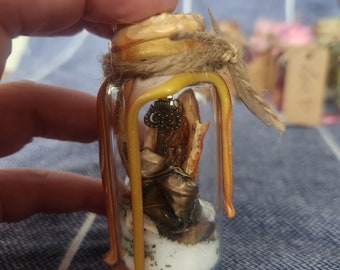 May include: A small glass bottle with a yellow wax seal and a brown twine bow. Inside the bottle is a small metal key, a piece of dried orange peel, and white sand.
