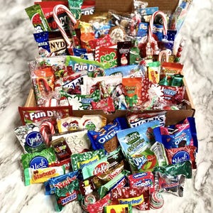 May include: A box filled with a variety of Christmas-themed candy and snacks, including chocolate bars, candy canes, and Fun Dip. The box is overflowing with treats, making it a perfect gift for any candy lover.