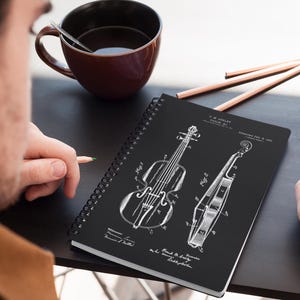 May include: A black spiral notebook featuring a white patent drawing of a violin. The patent drawing is titled "F.M. Ashley Violin" and "Patented Feb. 8, 1901".