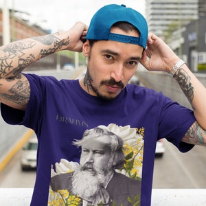 May include: A man with tattoos on his arms is wearing a purple t-shirt with a graphic of Johannes Brahms and the text "BRAHMS" printed on the shirt. He is also wearing a blue baseball cap.