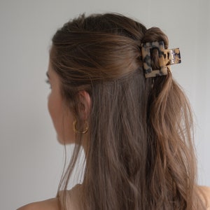May include: A tortoiseshell hair clip with a square shape, holding back a woman's long brown hair.