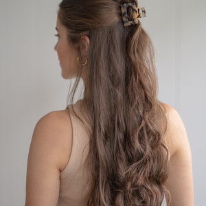 May include: A woman with long, wavy brown hair is wearing a beige tank top and black pants. She has a tortoiseshell hair clip in her hair.