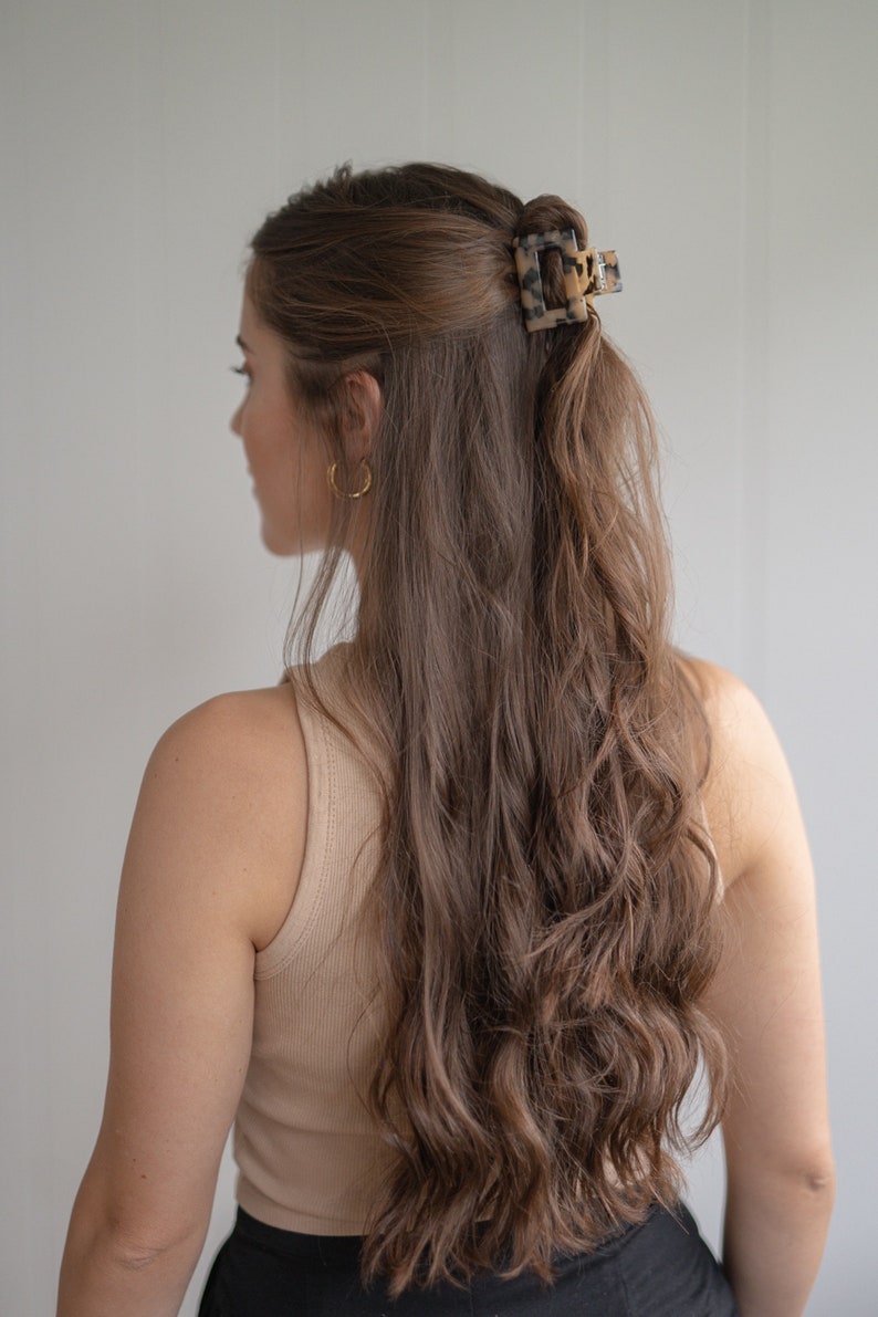 May include: A woman with long, wavy brown hair is wearing a beige tank top and black pants. She has a tortoiseshell hair clip in her hair.