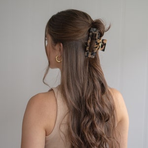 May include: A woman with long, wavy brown hair wearing a beige tank top and black pants. Her hair is pulled back with a large, tortoiseshell hair clip.
