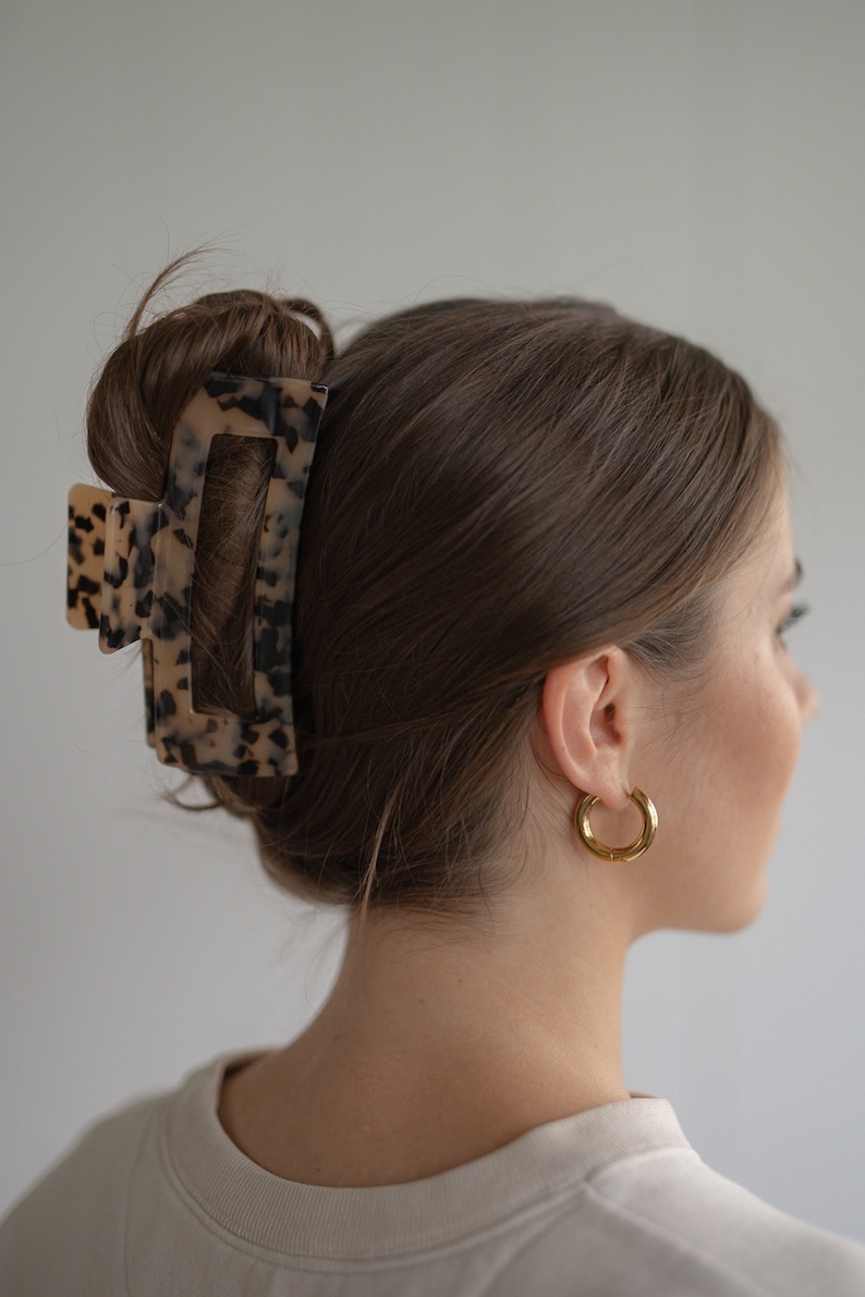 May include: A brown and black tortoiseshell hair claw with a rectangular shape. The claw is holding a woman's brown hair in a bun.