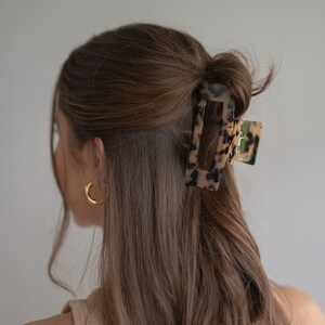 May include: A tortoiseshell hair clip with a rectangular shape and a smooth, glossy finish. The clip is holding a section of brown hair.