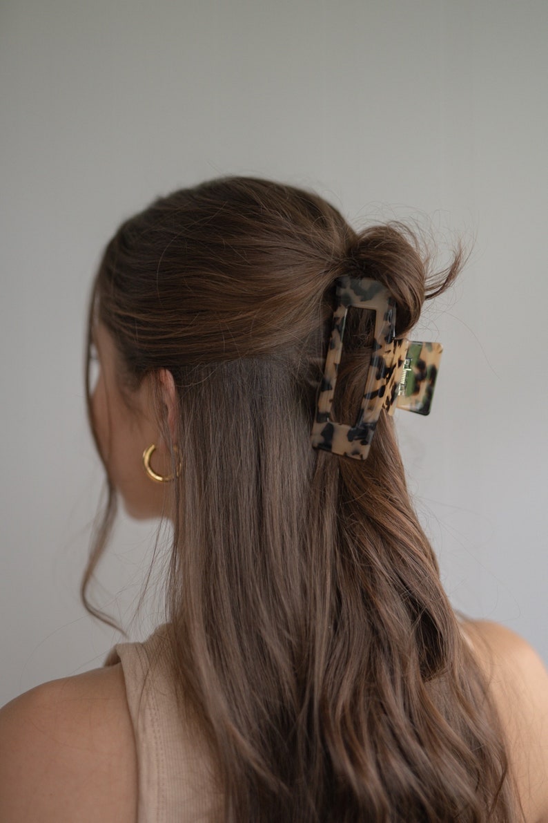 May include: A tortoiseshell hair clip with a rectangular shape and a smooth, glossy finish. The clip is holding a section of brown hair.