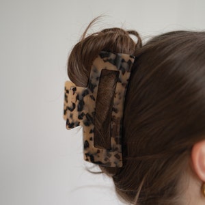 May include: A tortoiseshell hair claw with a rectangular shape and a smaller, triangular piece on the side. The claw is holding a bun of brown hair.