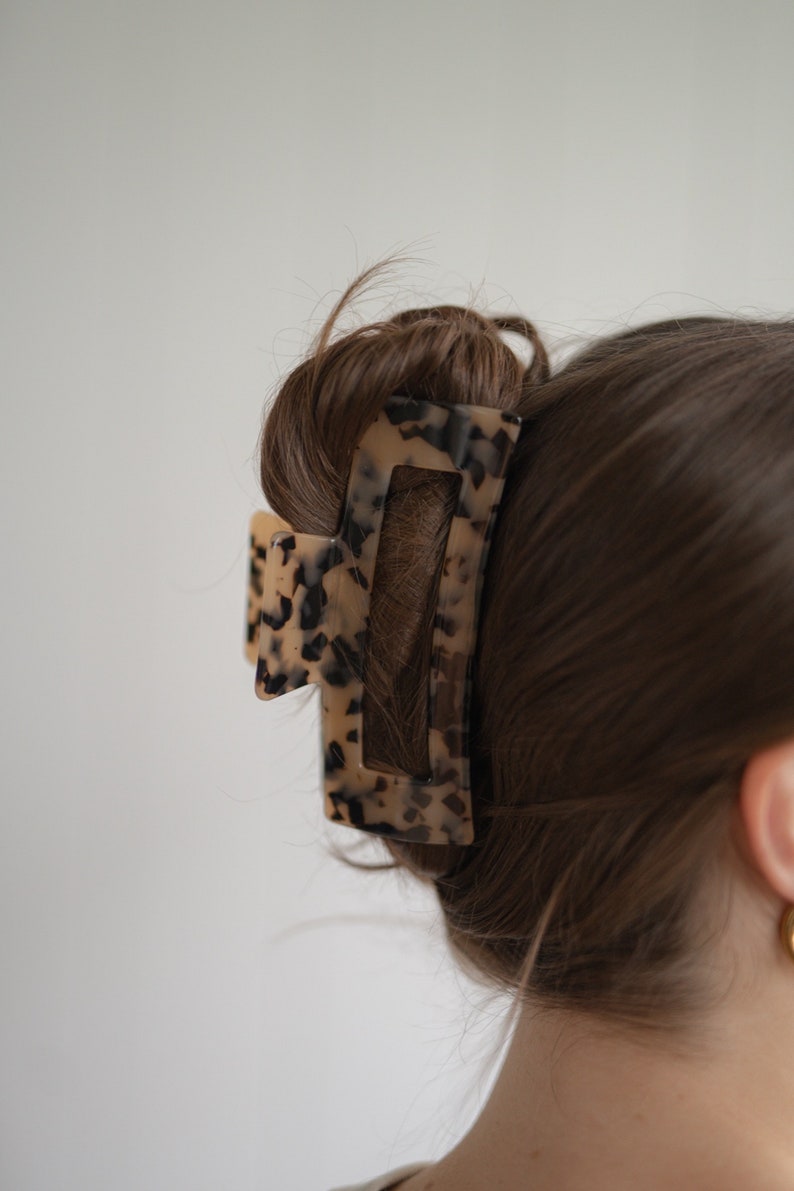 May include: A tortoiseshell hair claw with a rectangular shape and a smaller, triangular piece on the side. The claw is holding a bun of brown hair.