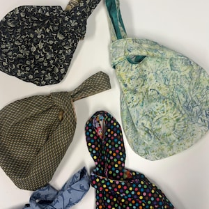 May include: Five fabric knot bags with various patterns including a black floral print, a light green abstract design, a dark olive geometric pattern, a light blue scroll print, and a black fabric with a colorful multi-colored polka dot pattern.