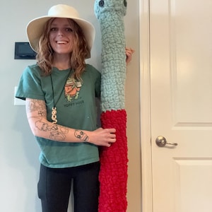 May include: A large, handmade crochet worm in shades of blue and red. The worm is held upright, showcasing its textured, plush design. The head is blue with two black button eyes. The body transitions to a vibrant red. A fun, whimsical home decor item.