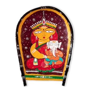May include: A hand-painted wooden fan depicting a mother goddess holding a baby elephant. The fan is decorated with intricate details and vibrant colors, including red, yellow, green, and white.