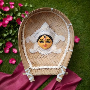 May include: A white and gold decorative face of a Hindu goddess in a woven bamboo basket. The face has a red dot on the forehead and a black bindi.