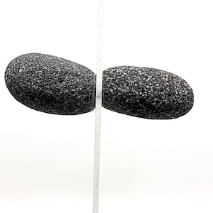 Op de afbeelding: Twee gladde, donkergrijze stenen met een ruw, getextureerd oppervlak zijn in evenwicht op een wit, verticaal oppervlak.