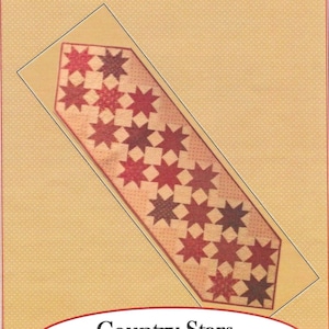 May include: A rectangular table runner with a country stars design. The runner is 18 inches wide and 70 inches long. The design features red and brown stars on a beige background. The text "Farmhouse Threads" is at the top.