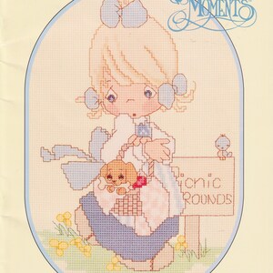 May include: A cross-stitch pattern booklet titled Precious Moments featuring a character with light hair and blue bows, holding a small brown puppy in a basket. The text reads Designs by Gloria & Pat, Precious Moments, and Picnic Rounds.