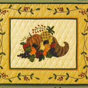 May include: Quilted rectangular placemat with a harvest theme. The center features a cornucopia overflowing with fruits and vegetables, including a pumpkin, pineapple, and grapes. The border is yellow with a diamond pattern and a vine design.