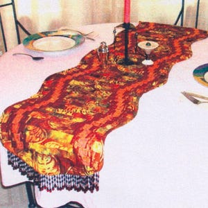 May include: A decorative table runner with a wavy design, featuring a mix of orange, red, and yellow patterns. The runner is adorned with a black beaded fringe. A white tablecloth and place settings are visible.