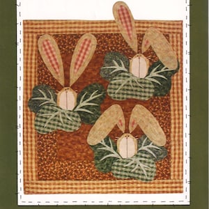 Può includere: Un arazzo da parete con tre figure di conigli con orecchie e facce beige, su uno sfondo a quadri marrone e verde. I conigli sono circondati da foglie verdi. L'arazzo da parete misura 61 cm per 74 cm.