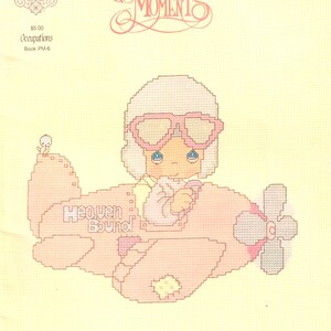 May include: A cross-stitch design featuring a cartoon pilot in a pink airplane with the words "Heaven Bound." The pilot wears pink goggles and a white helmet. The design is from the "Precious Moments" collection.