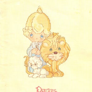 May include: Cross-stitch pattern featuring a blonde child with a lion and a small dog. The child wears a blue dress and holds a sprig. The lion is tan, and the dog is white. The words "Peace on Earth" and "Precious Moments" are visible.