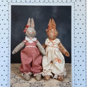May include: A pattern for making two fabric bunny dolls named "Amanda Marie". The dolls are dressed in vintage-style clothing. One doll is wearing a red and white dress with a white collar and the other is wearing a white dress with a floral pattern and a heart design.