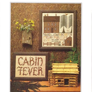 May include: An image featuring a cross-stitch pattern titled "CABIN FEVER." The design includes a framed cabin scene with a house, trees, and an alphabet. A small wooden cabin model and a framed text piece that reads "CABIN FEVER" are also present.