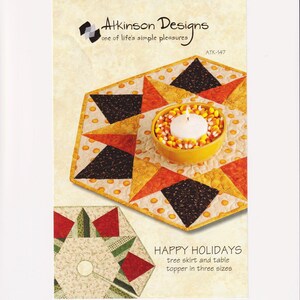 Könnte beinhalten: Ein farbenfroher, gesteppter Tischaufsatz mit einer Kerze in einer gelben Schale, umgeben von Zuckermais. Das Design zeigt orange, gelbe und schwarze Dreiecke. Der Text lautet "HAPPY HOLIDAYS" und "Baumrock und Tischaufsatz in drei Größen".