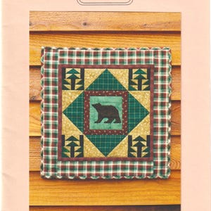 Könnte beinhalten: Eine gesteppte Wandbehänge mit einem Bärenmotiv. Der quadratische Quilt zeigt eine zentrale Bärensilhouette, umgeben von geometrischen Formen in Grün, Gold und Braun, umrahmt von einem karierten Rand. Die Maße betragen 43,8 cm x 43,8 cm.