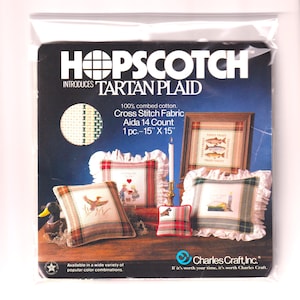 以下が含まれることがあります： 「HOPSCOTCH INTRODUCES TARTAN PLAID」のテキストが入ったクロスステッチキット。キットには、38cm x 38cmのAida 14カウント生地が含まれています。画像には、クッションや額装されたアートワークなど、完成したクロスステッチプロジェクトが表示されています。