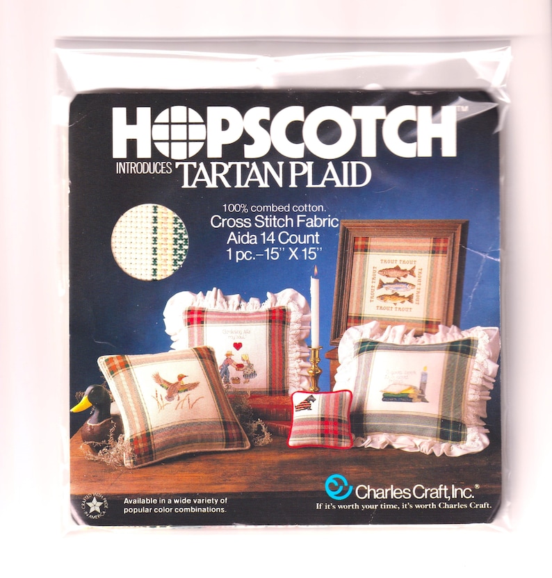 以下が含まれることがあります： 「HOPSCOTCH INTRODUCES TARTAN PLAID」のテキストが入ったクロスステッチキット。キットには、38cm x 38cmのAida 14カウント生地が含まれています。画像には、クッションや額装されたアートワークなど、完成したクロスステッチプロジェクトが表示されています。