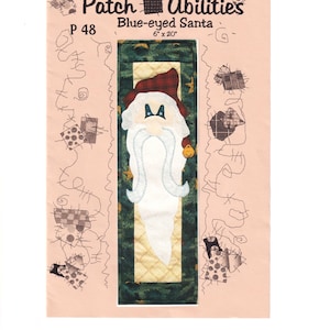May include: A craft pattern titled "Blue-eyed Santa" by Patch Abilities. The design features a Santa Claus with a white beard and a red plaid hat, set against a green and gold quilted background. The pattern measures 6" x 20".
