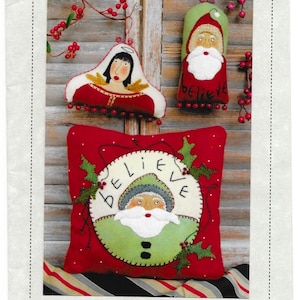 Peut inclure: Une présentation festive sur le thème de Noël avec un oreiller rouge et deux ornements. L'oreiller, mesurant 30 cm, présente un motif de Père Noël avec le mot "BELIEVE". Les ornements représentent une femme et le Père Noël. Le texte "Reets' Rags To Stitches" est également visible.