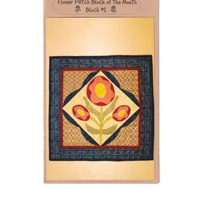 Puede incluir: Un bloque de tela enmarcado con un diseño floral. La imagen central muestra una flor grande y estilizada con pétalos rojos y amarillos, rodeada de flores más pequeñas. El bloque está bordeado por una tela azul oscuro con un diseño estampado. El texto "Flower Patch Block of the Month Block #1" está en la parte superior.