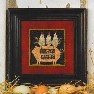 Könnte beinhalten: Gerahmte Punchneedle-Stickerei mit schwarzem Rahmen. Das Kunstwerk zeigt drei Hühner auf einem Schwein mit den Worten "Fresh Eggs" und der Zahl "2995". Der Text "THREADS THAT BIND" und "DESIGNER SHAWN WILLIAMS" stehen oben. Das Kunstwerk ist 11,4 cm x 11,4 cm groß.