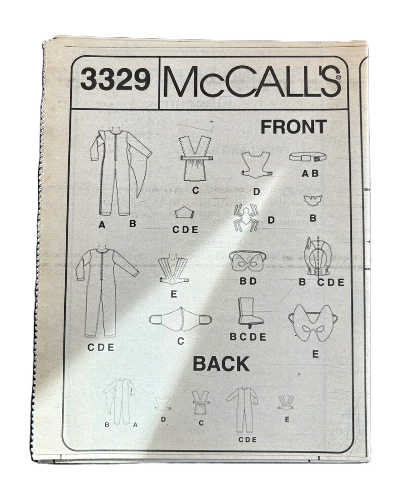 May include: A sewing pattern for a costume with the number 3329 and the brand name McCall's. The pattern includes instructions for making a jumpsuit, a cape, a mask, and a belt. The pattern is labeled "Front" and "Back" and includes different pieces labeled with letters.