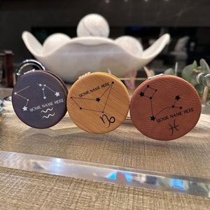 May include: A stack of personalized compact mirrors with various zodiac constellations. The mirrors have a silver rim and a leather-like cover in different colors. The text "YOUR NAME HERE" is printed on each mirror.