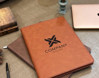 May include: A tan leather-effect portfolio folder with a zip closure, featuring a black logo with an X-shaped icon and the text COMPANY SLOGAN printed on the front cover, resting on top of a dark brown leather-effect folder and a black pen.