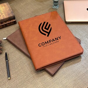 May include: A tan leather-like portfolio folder featuring a black abstract logo and the text COMPANY TAG LINE HERE printed on the front cover. The folder is resting on a darker brown leather-like surface next to a black pen and a laptop.